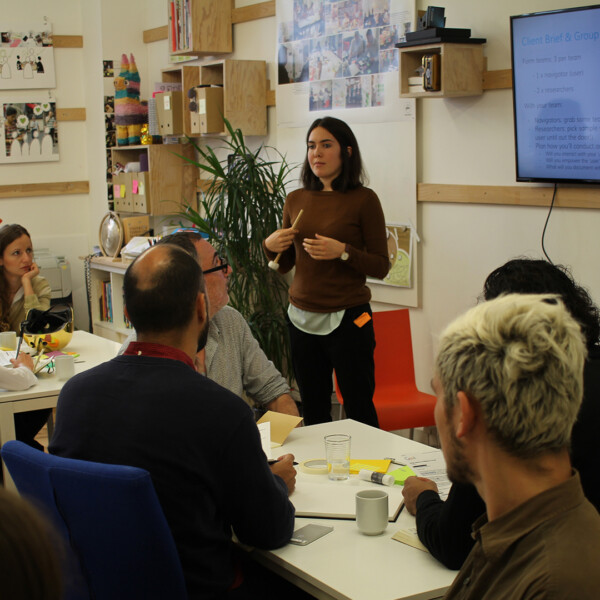 Woman presenting in workshop to group of people seated at tables.
