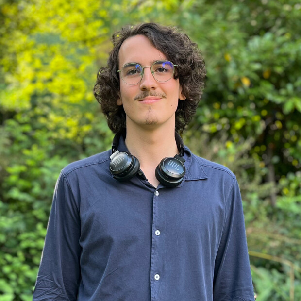 Outdoor photo of Yesse in a blue collared shirt with headphones around his neck, standing in front of a green bush.