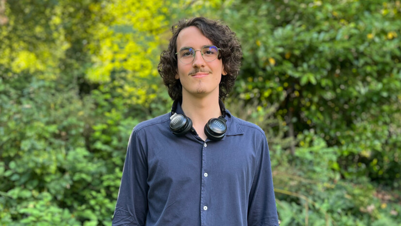 Outdoor photo of Yesse in a blue collared shirt with headphones around his neck, standing in front of a green bush.
